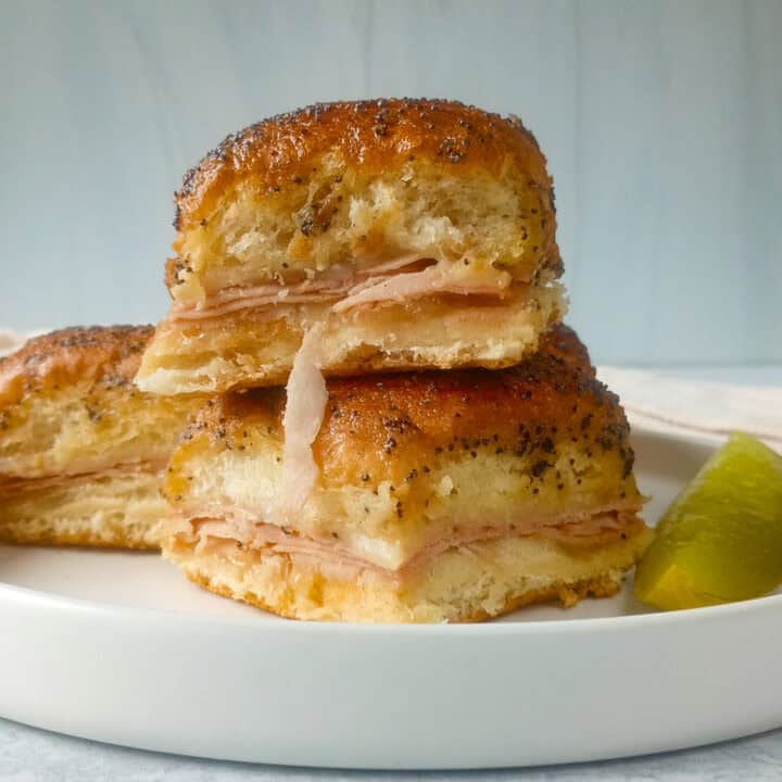 Picture of ham and Swiss Sliders stacked on a white plate next to a pickle.