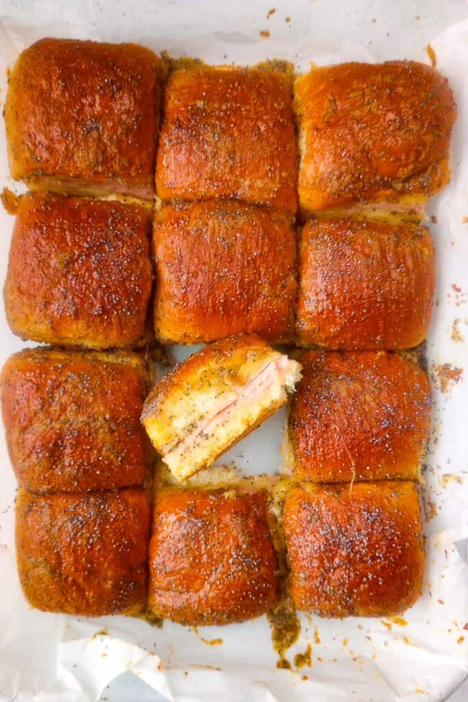 Overhead picture of Baked ham and cheese sliders fresh out of the oven with glaze sauce. One slider in the middle is flipped on its side, showing the inside of the sandwich. 