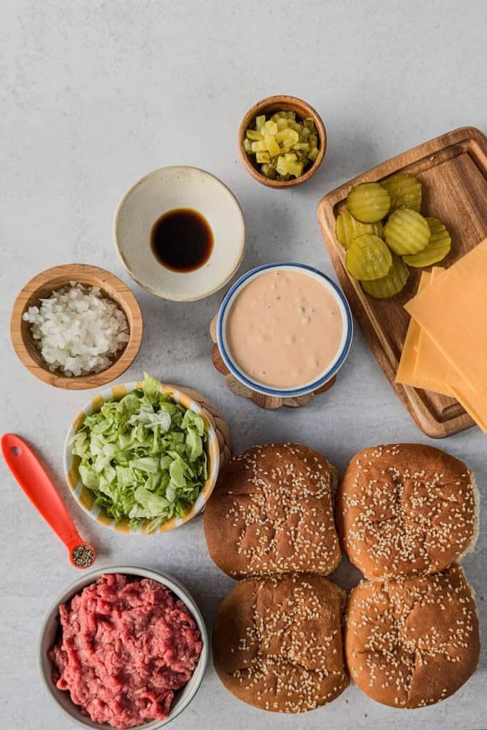 Overhead picture of ground beef, coarse black pepper, Worcestershire sauce, diced onion, diced pickles, dill hamburger pickle chips, American cheese, sesame seed buns, shredded lettuce, and big Mac sauce. 