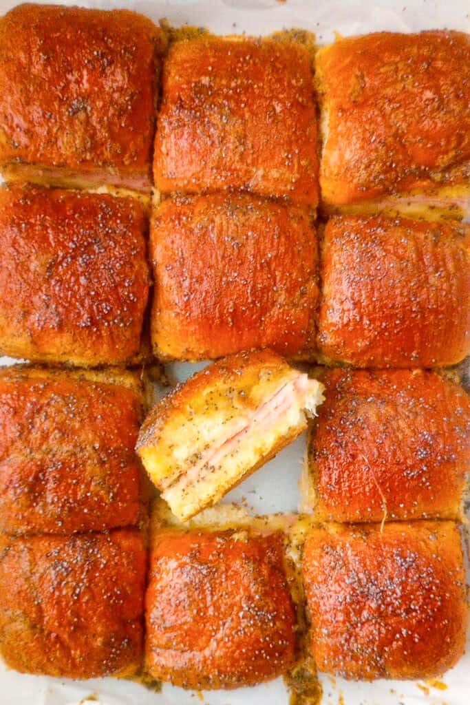 A tray of Hawaiian roll slider sandwiches fresh out of the oven, with one tipped on its side so you can see the ham and melted cheese inside.