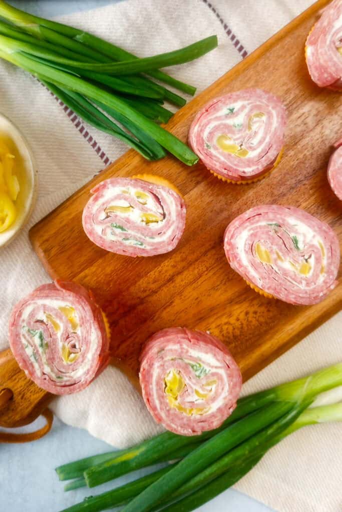 Salami rolls cut into into pinwheel shape on crackers. Green onions and banana peppers are arranged on the side. 