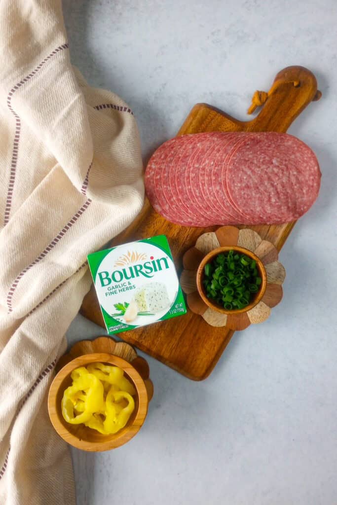 Overhead picture of ingredients needed to make salami and cream cheese roll ups. Hard dry salami, Boursin garlic and fine herbs cheese spread, diced green onions and banana peppers. 