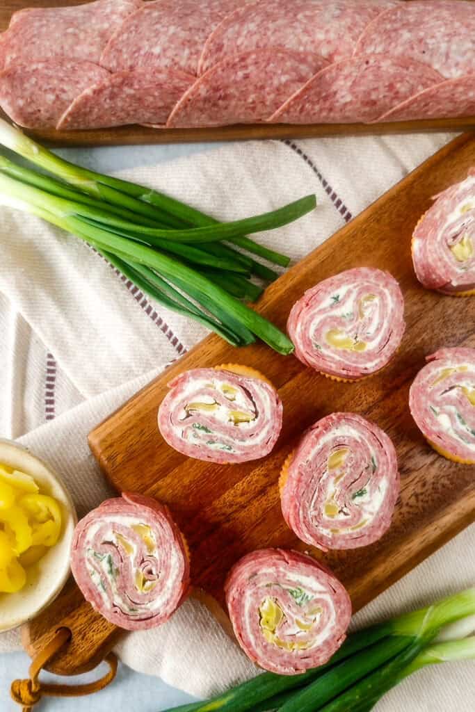 Picture of salami pinwheels on a brown cutting board next to a bowl of banana peppers and green onions. Another, rolled Salami log is arranged in the background. 