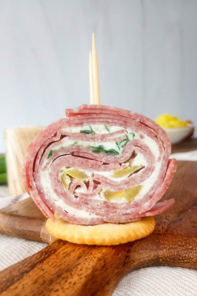 Up close picture of salami pinwheel on a cracker with a toothpick inserted in the top. Bowl of banana peppers arranged in the background along with green onions and a clear bowl of toothpicks.