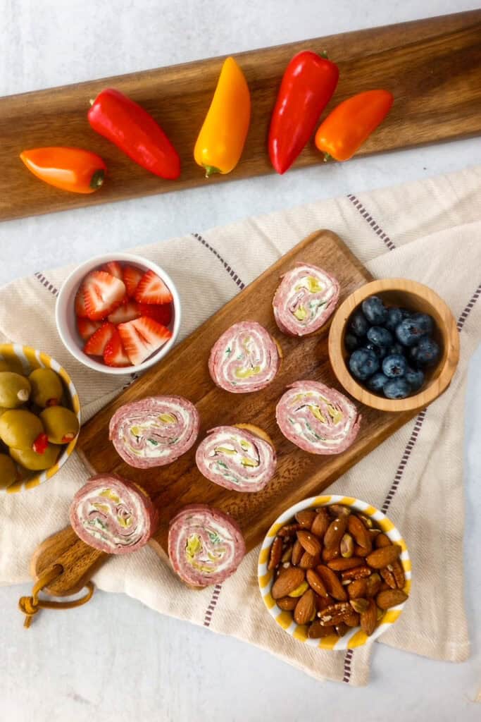 Overhead picture of sliced salami cream cheese roll ups on a brown cutting board. In small bowls, there are charcuterie board items; such as, sliced strawberries, blueberries, green olives, and mixed nuts. 