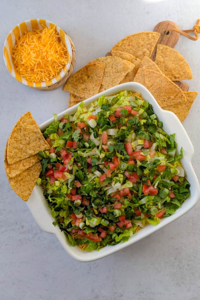 Overhead picture of Mexican taco dip ready to eat arranged next to a bowl of shredded cheddar cheese and tortilla chips. 