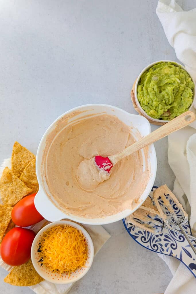 Overhead picture of Roma tomatoes, tortilla chips, guacamole, shredded cheese, and blended cream cheese and sour cream seasoned with taco seasoning. 