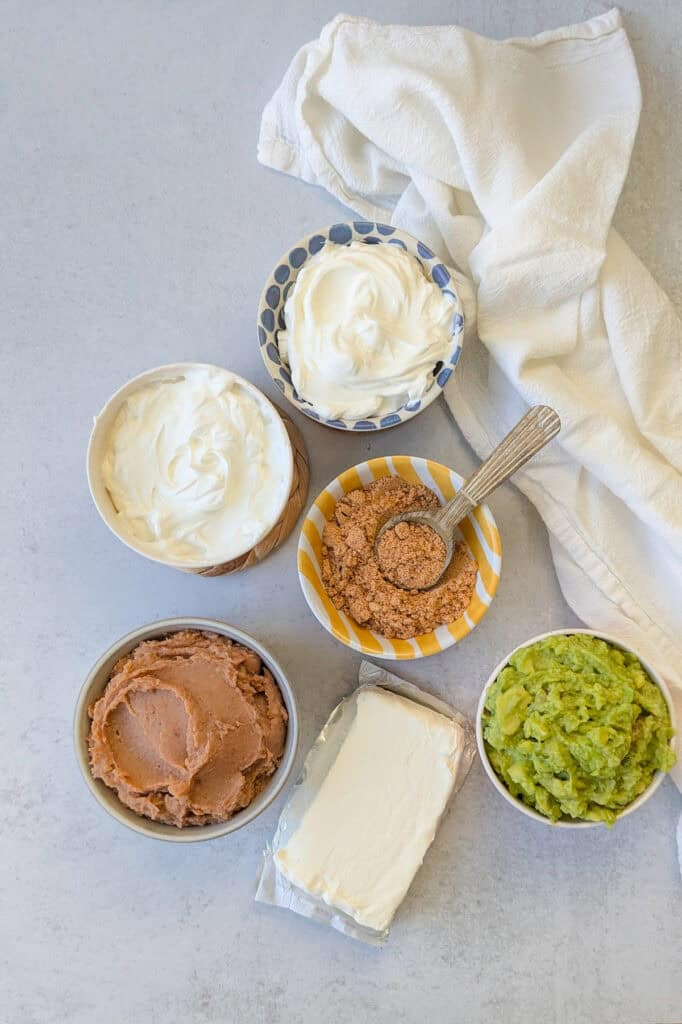 Overhead picture of ingredients needed to make layered taco dip; refried beans, cream cheese, sour cream, taco seasoning, and guacamole. 