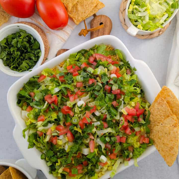 Overhead picture of taco dip next to shredded lettuce, diced tomatoes, chopped cilantro and tortilla chips.