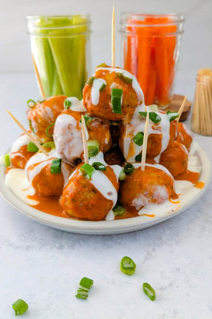 Picture of meatballs on a plate slathered in buffalo sauce, drizzled with ranch dressing, and garnished with chives. Carrot and celery sticks are arranged in glass jars in the background and toothpicks are set in chicken meatballs for dimension.