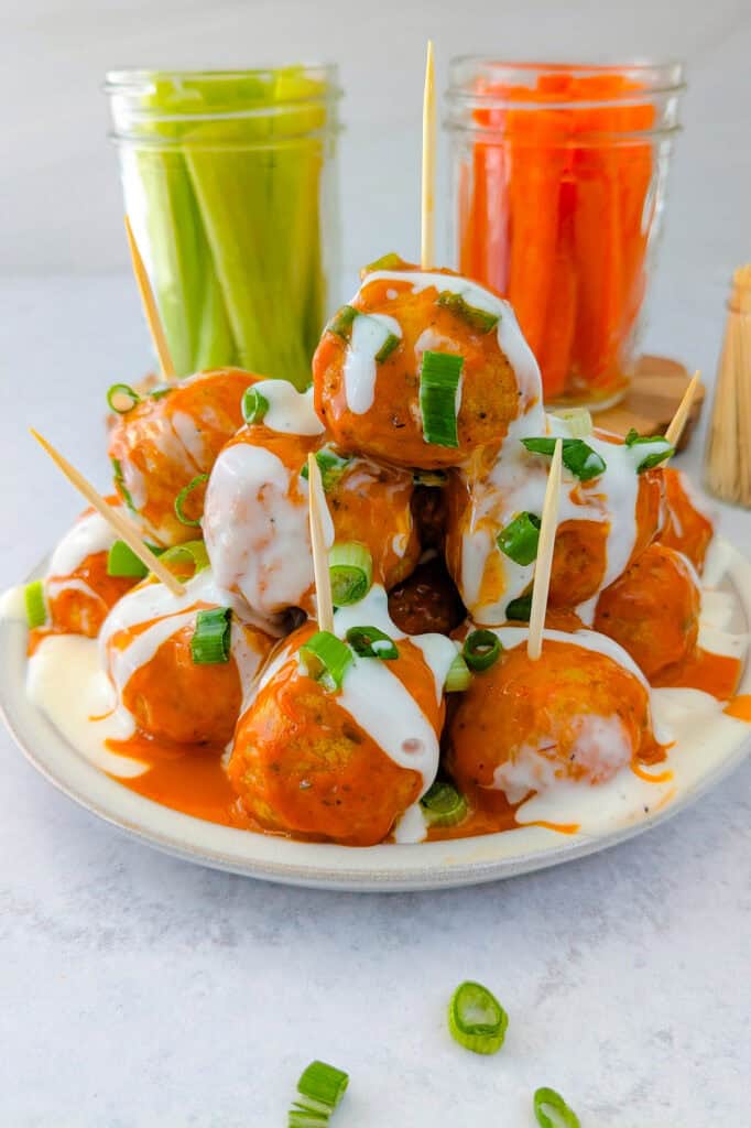 picture of meatballs on a plate garnished with chives, slathered in creamy buffalo sauce and drizzled with buttermilk ranch dressing. Carrot and celery sticks are arranged in the background in glass jars. 