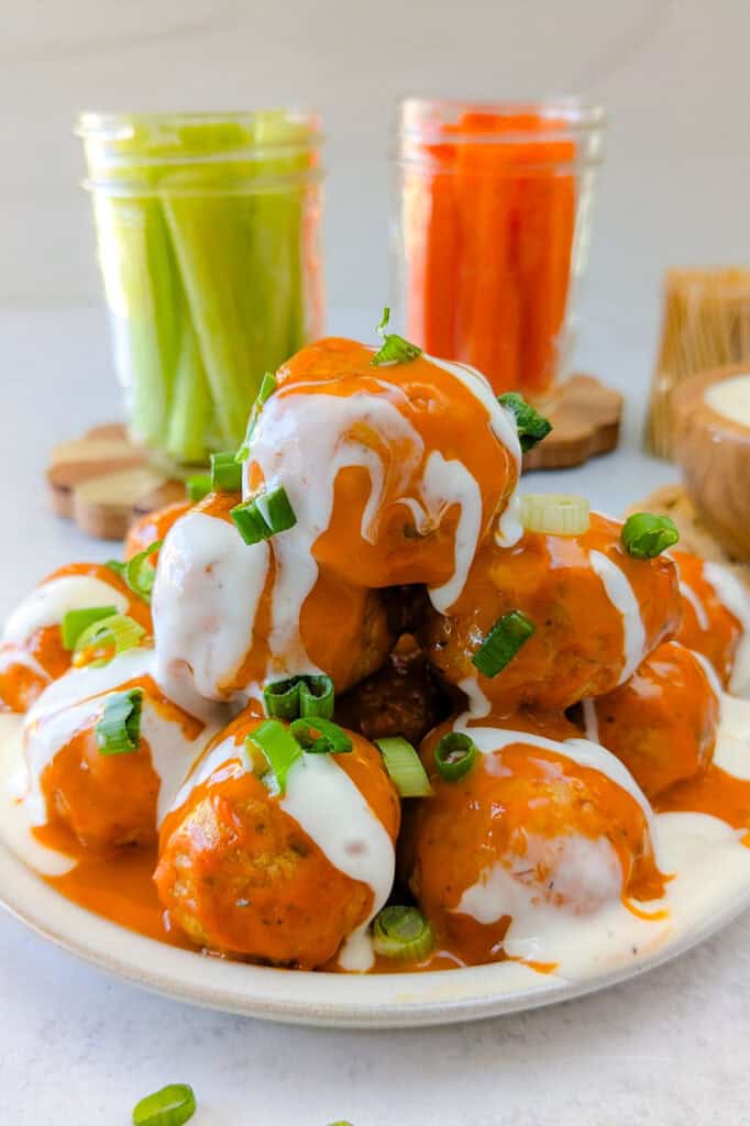 Picture of crock-pot buffalo chicken meatballs on a plate slathered in spicy buffalo sauce, drizzled with ranch dressing, and garnished with chives. 