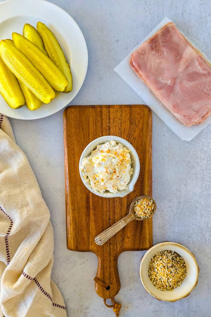 Overhead ingredients of pickle spears, deli ham, cream cheese, and everything bagel seasoning. 