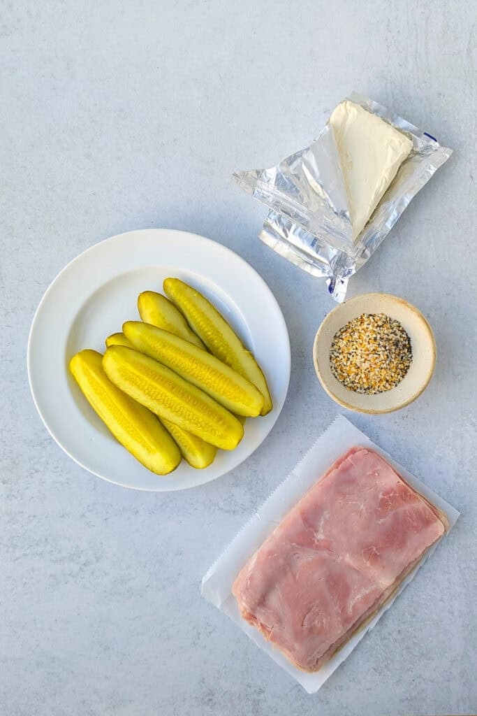 Overhead picture of dill pickle spears, deli ham, cream cheese, and everything bagel seasoning. 