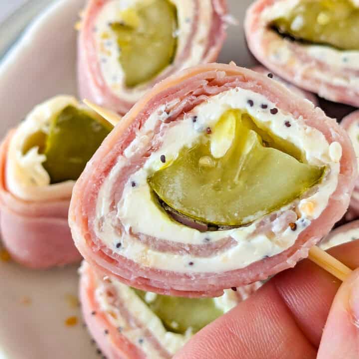 Up close picture of dill pickle roll up on a toothpick angled over a plate with sliced pickle pinwheels.