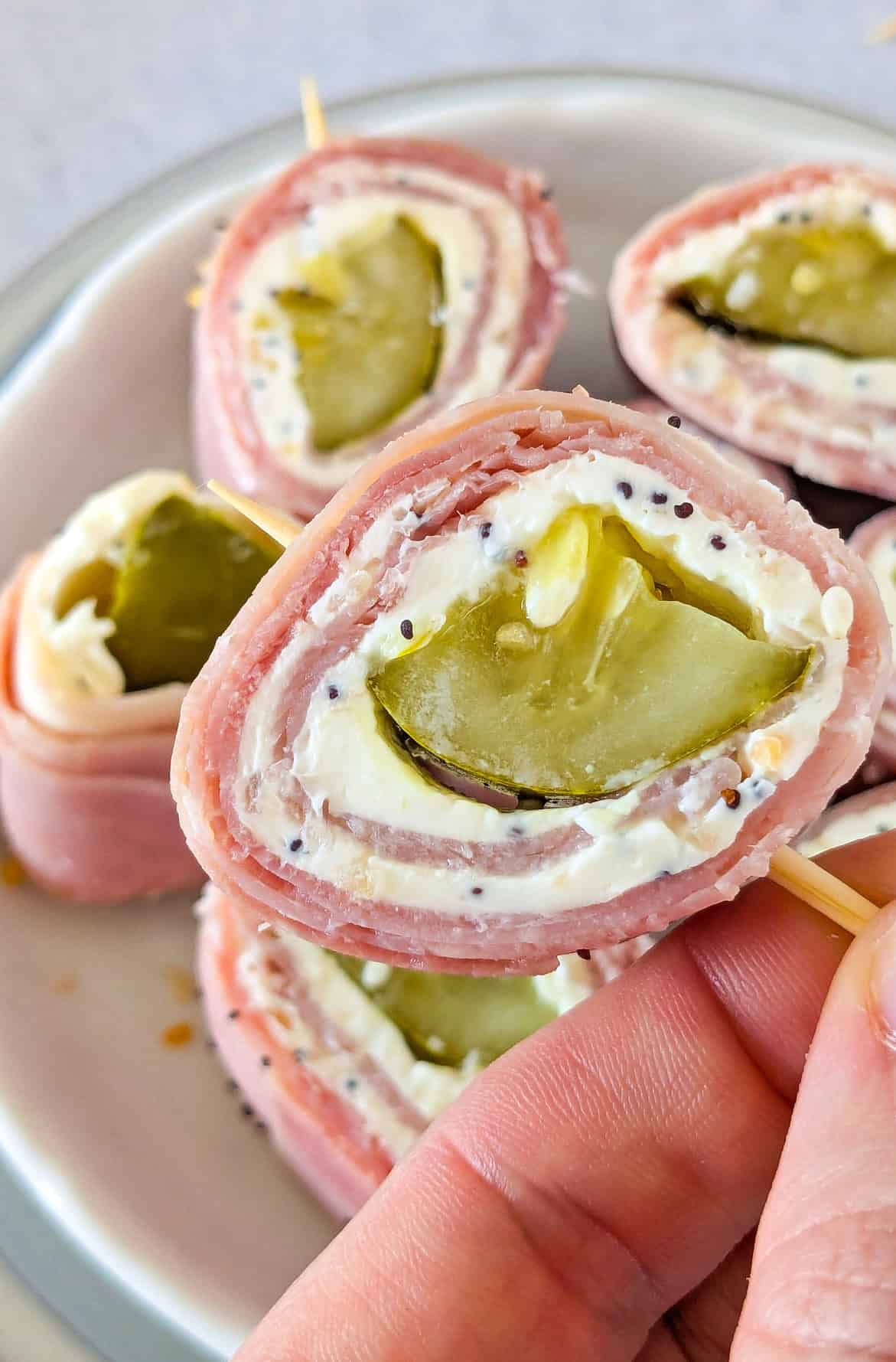 Up close picture of dill pickle roll up on a toothpick angled over a plate with sliced pickle pinwheels.