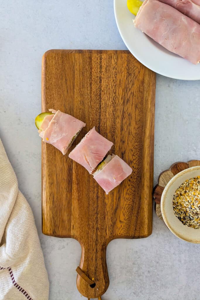 Overhead picture of sliced pickle wrap on a brown chopping board. More pickle wraps are on a white plate in the upper right corner and everything bagel seasoning is in a bowl on the right side of the chopping board. 