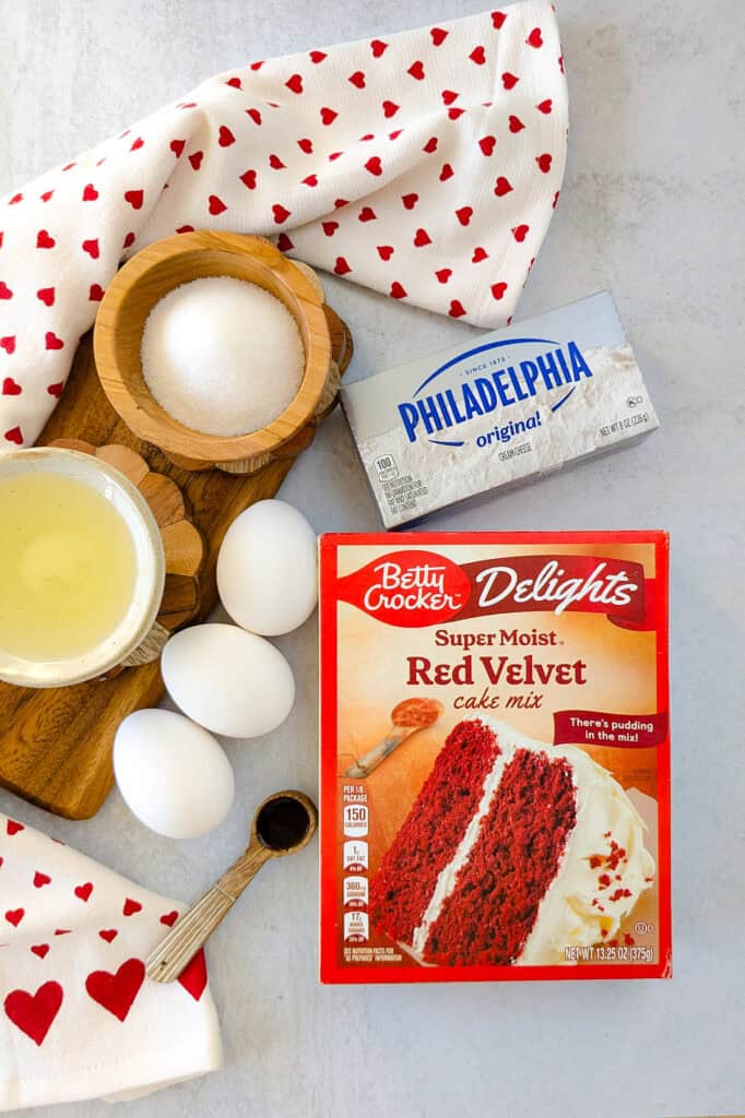 Overhead picture of ingredients for red velvet cheesecake brownies. Betty Crocker red velvet cake mix, cream cheese, eggs, oil, sugar, vanilla extract. 