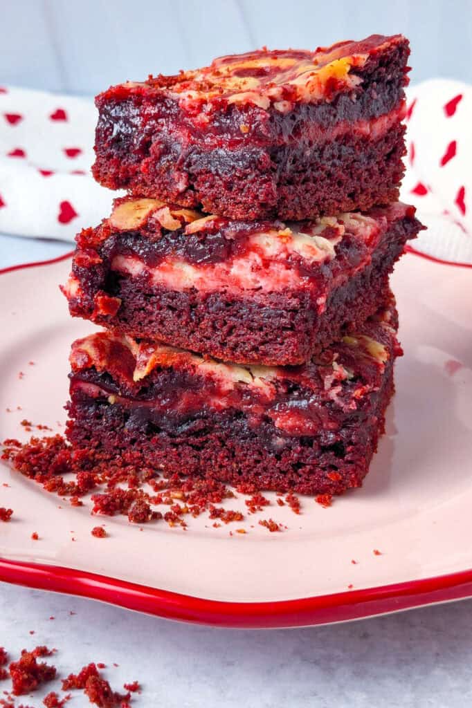 Up close picture of brownies stacked on a red and pink plate. A white towel with hearts is layered in the background. 