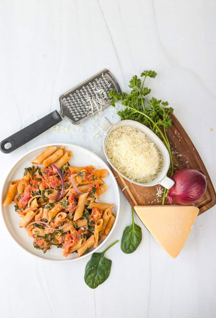 Overhead picture of baked pasta on a white plate next to grated cheese, parsley, red onion, and spinach. 