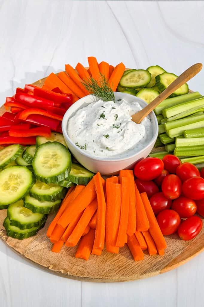 Picture of healthy Greek yogurt dip in a white bowl with fresh dill and a wooden spoon. Vegetables are angled all around.