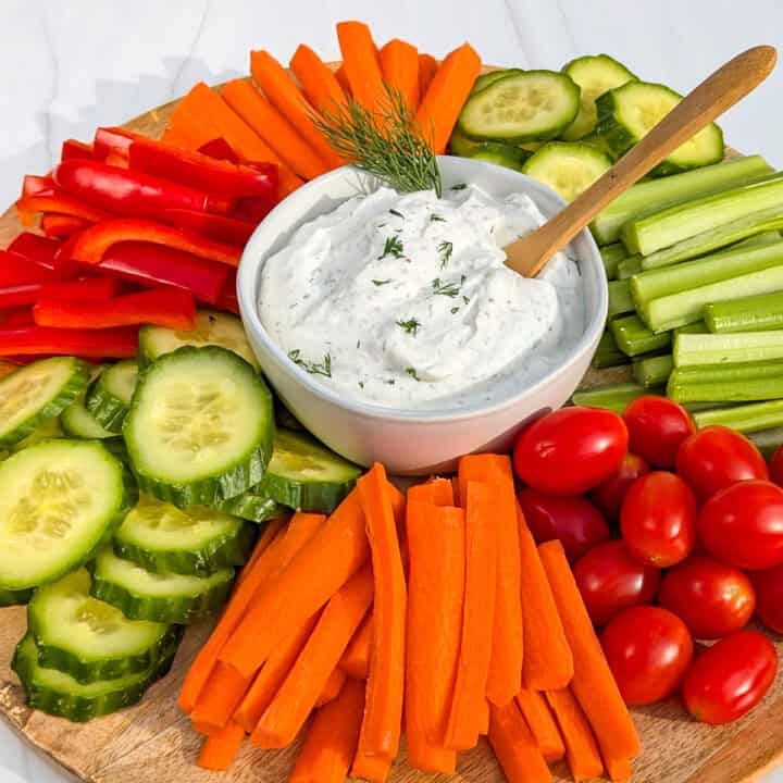 Picture of healthy Greek yogurt dip in a white bowl with fresh dill and a wooden spoon. Vegetables are angled all around.