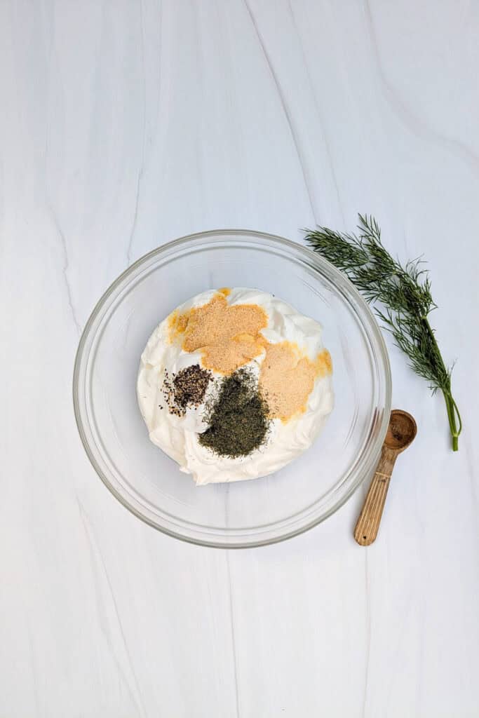 Overhead picture of Greek yogurt in a bowl with garlic powder, dill weed, onion powder, salt, coarse black pepper, and organic apple cider vinegar. 