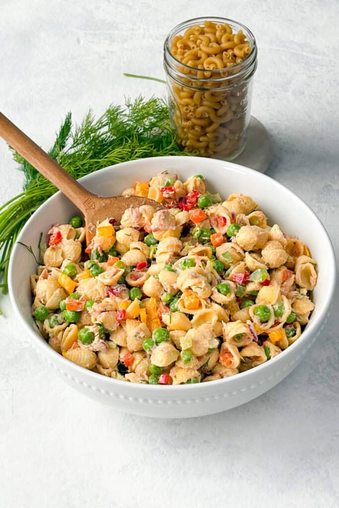 Tuna pasta salad in a bowl with a wooden serving spoon. Noodles and fresh dill are arranged in the background.