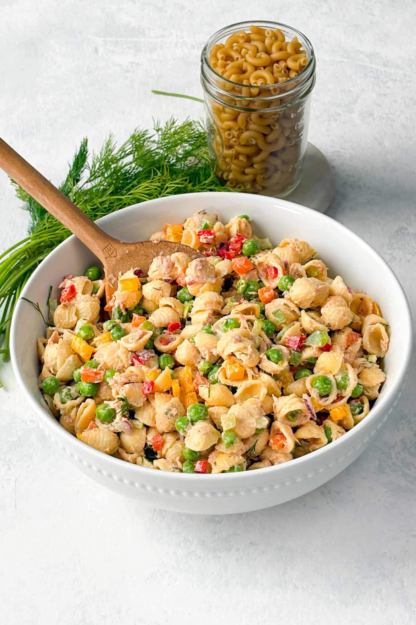 Tuna pasta salad in a bowl with a wooden serving spoon. Noodles and fresh dill are arranged in the background.
