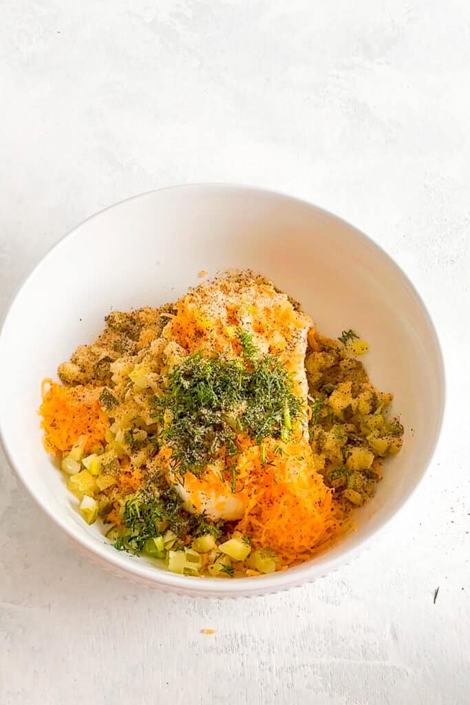 Overhead picture of cream cheese, diced dill pickles, pickle juice, fresh dill, and shredded cheddar cheese in a large white bowl.