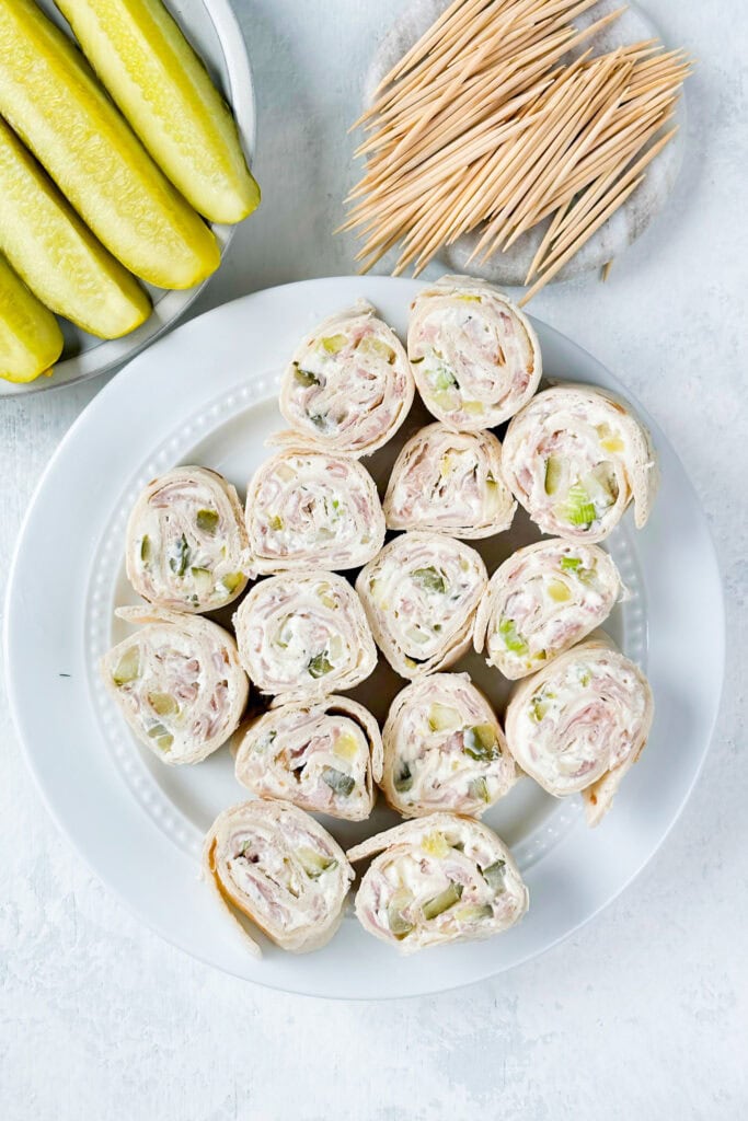 Overhead picture of pickle dip pinwheels on a white plate next to pickle spears and toothpicks.