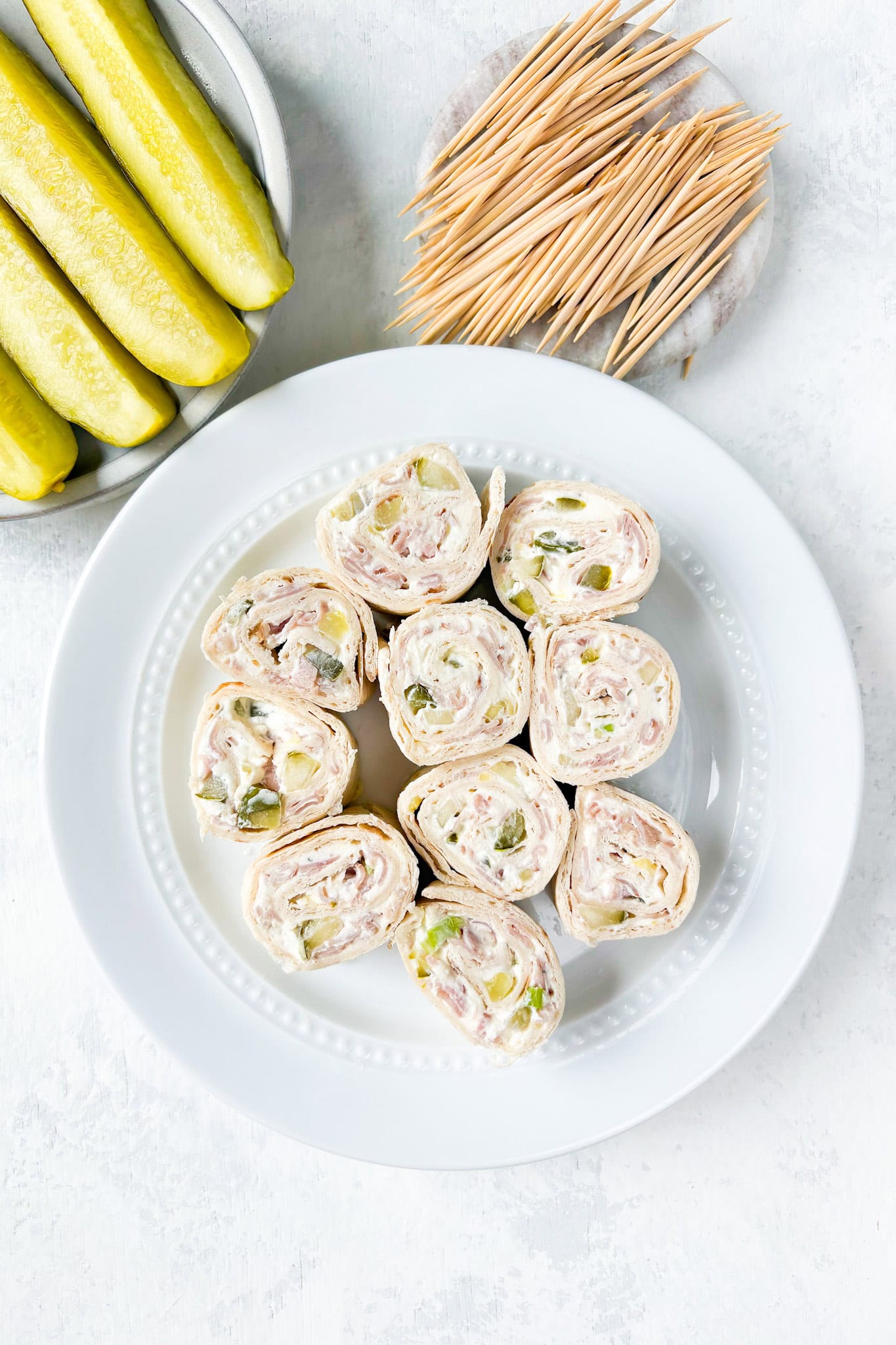 Overhead picture of pickle dip pinwheels on a white plate next to pickle spears and tooth picks.