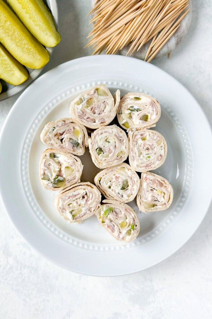 Overhead picture of pickle roll ups on a white plate next to dill pickle spears and toothpicks.