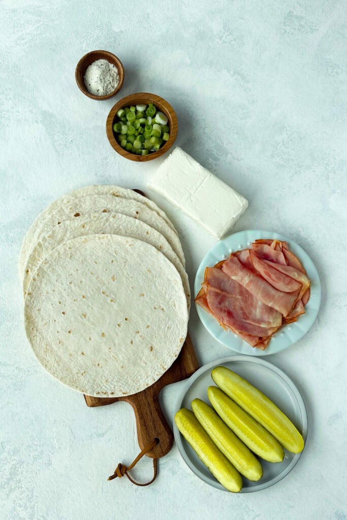 Overhead picture of ingredients; cream cheese, dill pickles, ham, green onions, ranch seasoning, and (8-inch) flour tortillas.