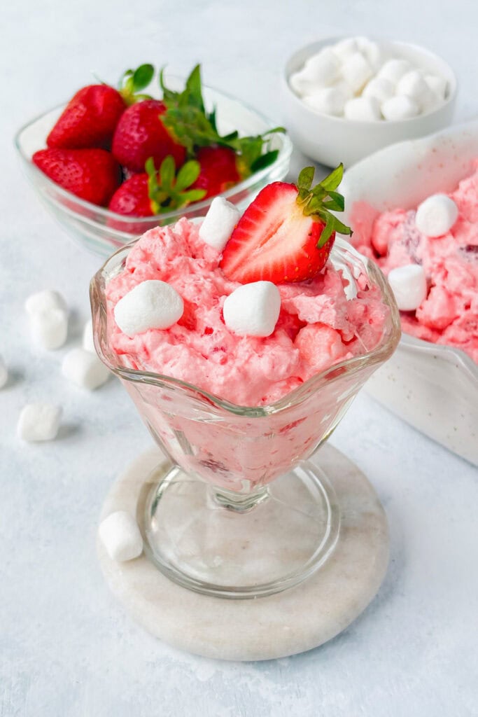 Strawberry Jello Flufif n a glass bowl garnished with a strawberry. A serving dish of of pink fluff, a small bowl of mini marshmallows, and a bowl of strawberries are in the background.