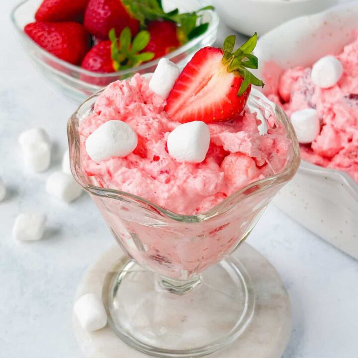 Strawberry Jello Flufif n a glass bowl garnished with a strawberry. A serving dish of of pink fluff, a small bowl of mini marshmallows, and a bowl of strawberries are in the background.