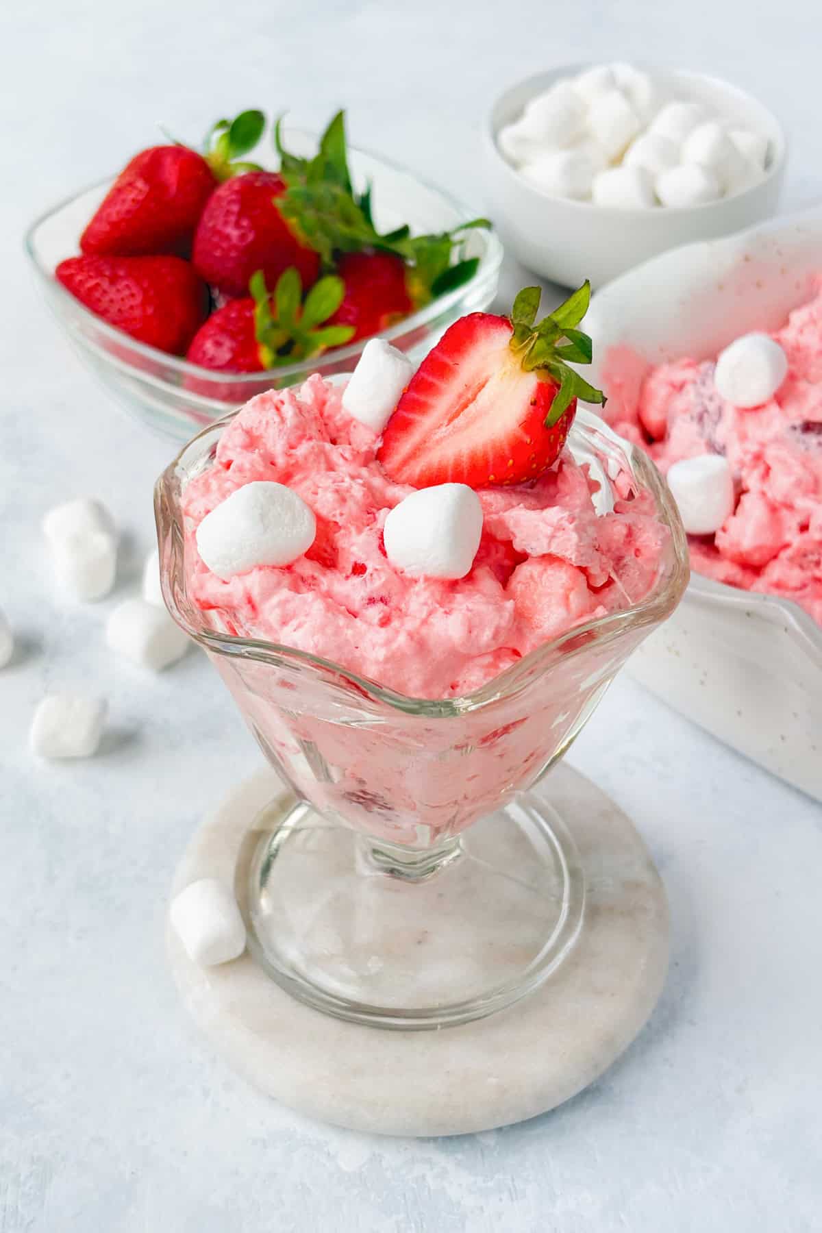 Strawberry Jello Flufif n a glass bowl garnished with a strawberry. A serving dish of of pink fluff, a small bowl of mini marshmallows, and a bowl of strawberries are in the background.